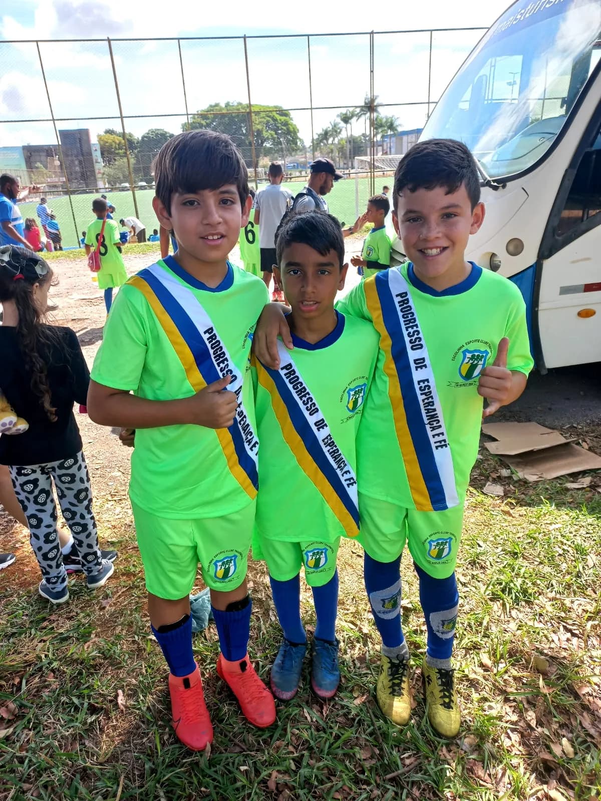 Três alunos de uniforme verde-limão com faixas comemorativas em frente ao ônibus da viagem